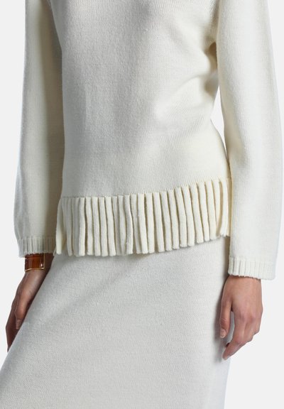Femme portant un pull en maille crème avec un ourlet côtelé et une jupe assortie, montrant le poignet avec une montre en cuir marron.