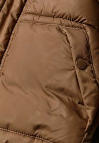 Tessuto di giacca a vento marrone con texture cucita, dotata di una grande tasca inclinata e di un accento con bottone rotondo marchiato. Finitura lucida e morbida.