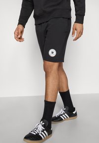 Mustat puuvillashortsit, joissa on istuva leikkaus ja vasemmassa lahkeessa merkki. Päällä mustat urheilusukat ja mustat lenkkarit.