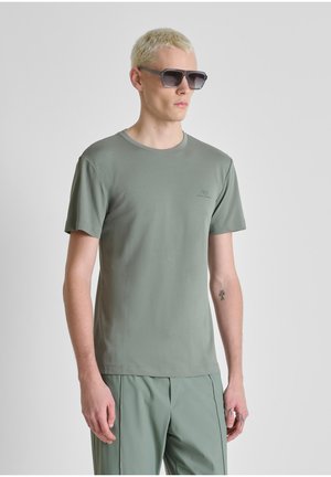 Uomo biondo che indossa occhiali da sole scuri oversize, t-shirt a maniche corte verde chiaro e pantaloni abbinati, in piedi davanti a uno sfondo semplice.