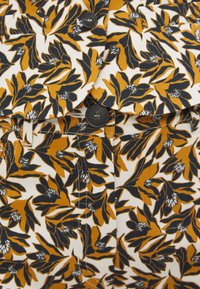 Tissu à motif floral présentant des feuilles noires et jaunes sur un fond blanc, avec des boutons noirs et des coutures détaillées.