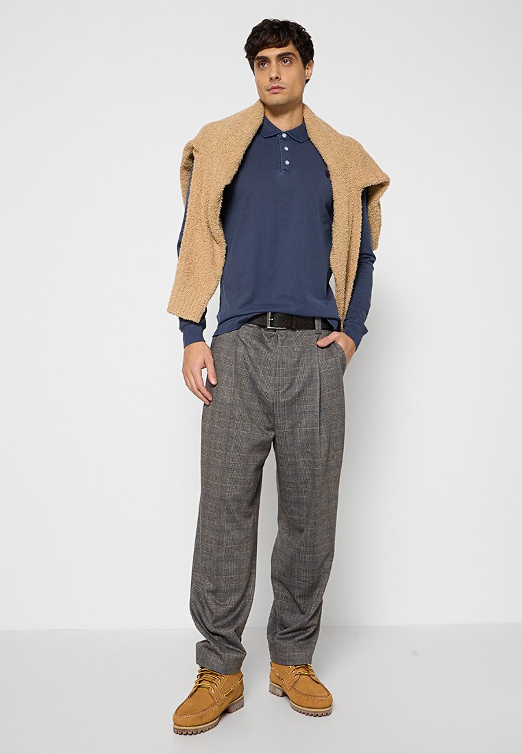 Marineblaues Langarm-Polo, beiger strukturierter Pullover über die Schultern gehängt, graue Karohosen und beige Schnürstiefel. Einfaches, lässiges Outfit.