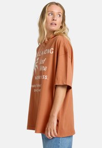 Camiseta de gran tamaño en marrón con texto gráfico en blanco y diseños. Hecha de material suave, con mangas cortas y un ajuste relajado.