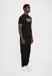 Sort bomulds t-shirt med EA7-logo, parret med sorte sweatpants. Modellen har hvide sneakers på og står i profil mod en ensfarvet baggrund.