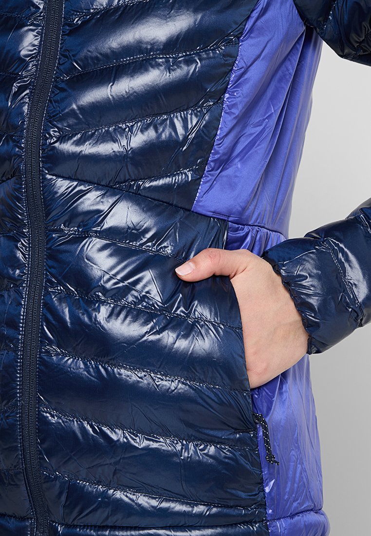 Doudoune bleu marine avec une texture brillante et matelassée. Présente un panneau latéral en contraste de couleur violet clair et une fermeture éclair à l'avant. Main dans la poche.
