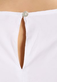 Blouse blanche avec une découpe en forme de goutte d'eau au dos, fermée par un bouton rond unique, confectionnée dans un tissu léger et texturé.