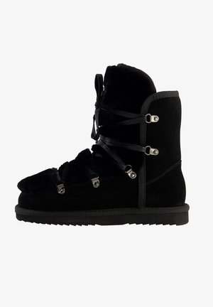Schwarzer Wildlederstiefel mit Kunstfellfutter, ausgestattet mit einer Schnürung an der Vorderseite, metallischen Akzenten und einer flachen Gummisohle. Design in knöchelhöhe.