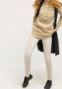 Camisa beige con cuello, de manga larga; jeans skinny blancos; cárdigan negro sobre los hombros; zapatos planos negros; textura suave, ajuste casual.