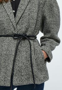 Grå og hvid frakke med kærnebenmønster, bælte med en sort snor, brede ærmer og forlommer. Teksturret uldmateriale.