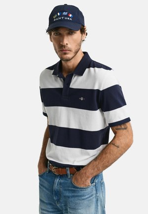 Marineblå og hvid stribet polo T-shirt med krave, to knapper og et lille logo. Kombineret med blå denim jeans og en marineblå kasket med tekst og flag.