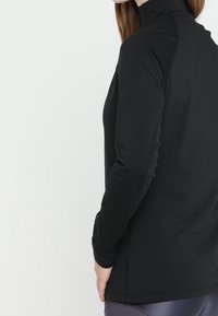 Mustapaitainen pitkähihaisvaate, jossa on korkea kaula-aukko, tiukka malli ja saumaton rakenne, valmistettu pehmeästä ja joustavasta kankaasta.