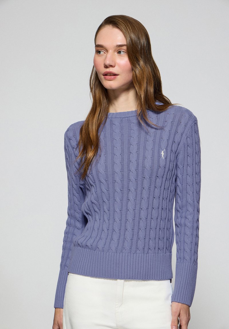 Lila kabelstickad tröja med ribbad mudd och nederkant, som har en liten, kontrasterande logotyp på bröstet. Mjuk textur, klassisk passform.