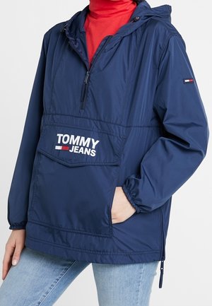 Granatowa wiatrówka Tommy Jeans z przednią kieszenią i półzamkiem, noszona na czerwonym golfie i jasnoniebieskich dżinsach.