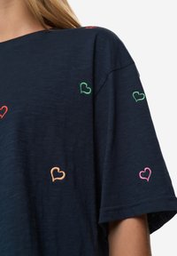 Camiseta azul marino de tela suave con diseños de corazones multicolores bordados en rojo, verde, rosa y naranja dispersos por el frente.