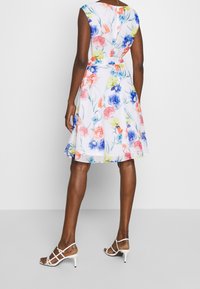 Kleid mit floralem Muster, tailliertem Schnitt, schwingendem Rock und ärmellosem Design. Die Farben umfassen Weiß, Rosa, Blau und Gelb. Kombiniert mit weißen Riemchenschuhen.