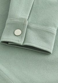 Collar de tela verde claro con una textura suave y detalle de botón plateado. Las costuras están cosidas y son visibles un borde limpio y doblado.