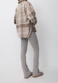 Przesadny beżowo-brązowy flanelowy kosz shirt w kratę, zestawiony z jasnoszarymi prążkowanymi spodniami dzwonowymi oraz szarymi butami typu slip-on z teksturowaną podeszwą.