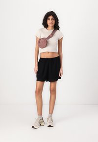Top corto in tessuto beige, pantaloni shorts neri con orlo in pizzo, e una borsa a tracolla rosa. Sneakers grigie chiare con dettagli scuri.