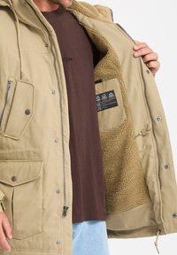 Khaki outer jacket with a zipper and snap closures, fleece lining, and multiple pockets. Interior features a care label on a black fabric patch.