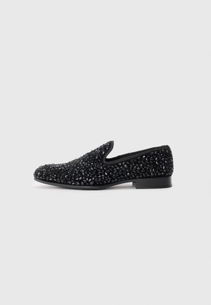 Svarte slips-on flate sko dekket med blanke svarte perler med lav hæl, vist i profil mot en hvit bakgrunn.