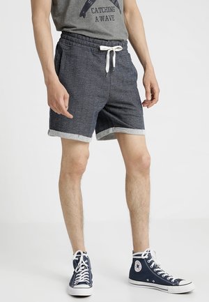 Mężczyzna noszący granatowe szorty z podwiniętymi nogawkami i białym sznurowadłem, szarą koszulkę z grafiką oraz granatowe wysokie trampki z białymi sznurowadłami.