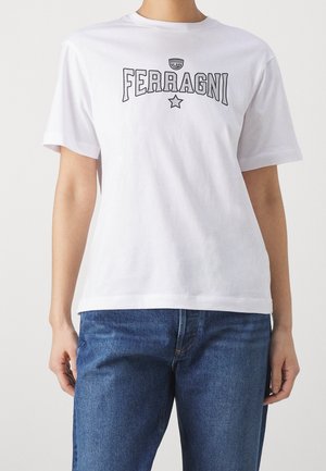 Personne portant un T-shirt blanc à manches courtes avec le mot "FERRAGNI", un logo œil, et une étoile imprimés sur la poitrine, assorti à un jean bleu.