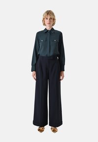Dunkelgrüne Bluse mit langen Ärmeln und zwei Brusttaschen, kombiniert mit weit geschnittenen, hoch taillierten navyblauen Hosen und goldenen Schuhen.