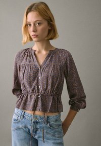 Langærmet, mønstret bluse i mørkeblå med blomster motiv, med v-hals, knapfront og snøre i kanten, parret med lys blå jeans.