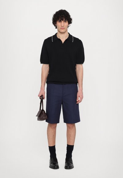 Polo-shirt en tricot noir avec un col blanc, shorts navy et chaussures noires. Le modèle tient un petit sac marron foncé. Design simple et moderne.