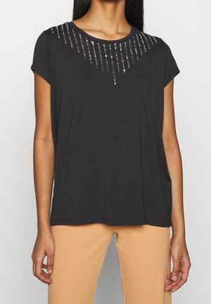 Frau trägt ein schwarzes kurzärmeliges Oberteil mit silbernen Pailletten vertikal entlang des Halsausschnitts und beigefarbene Hose.