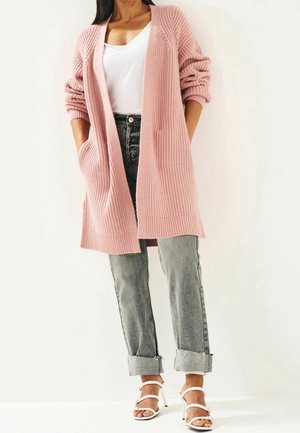 Hellrosa gestrickte Strickjacke mit lockerem Design, rollierten Ärmeln und Seitentaschen, kombiniert mit hochgeschnittenen grauen Jeans und weißen Sandalen.