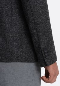 Manche de blazer gris texturé avec trois boutons noirs, montrant le détail de tissage fin. Porté par-dessus un tissu léger à motifs, mettant en évidence le contraste.