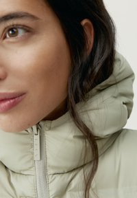 Ljusgrön pufferjacka med hög krage och mjuk, slät textur. Har en central dragkedjestängning och en lätt lyster.