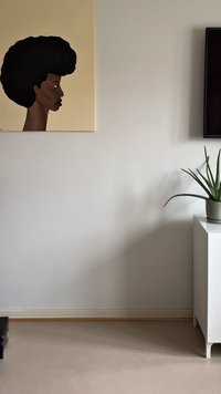 Vägg med en sidoprofilmålning av en svart kvinna med afrohår, vit skåp som håller en krukad aloe vera-växt, och en del av en monterad tv.