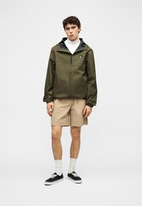 Νέος άνδρας που στέκεται, φορούσε χακί μπουφάν με φερμουάρ, μπεζ σορτς, λευκές κάλτσες και μαύρα αθλητικά παπούτσια, μπροστά σε απλό ανοιχτό φόντο.