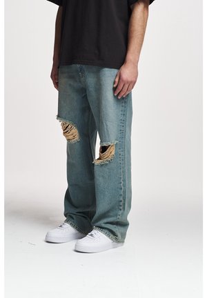Helle blaue Denim-Jeans mit ausgefransten Kniefen, rohen Kanten und einem lockeren Schnitt, kombiniert mit weißen Sneakers.