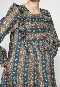 Šaty s květinovým potiskem v tmavě modrozelené barvě s volánkovým výstřihem a rukávy. Obsahují růžové a zelené vzory, textura látky a vrstvený lem.