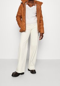 Giacca imbottita marrone con cappuccio, maglione in morbido lavorato a trecce color crema, pantaloni bianchi larghi e scarpe con zeppa marroni con suola testurizzata.