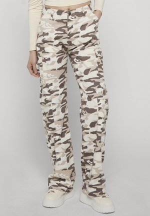 Persona che indossa pantaloni cargo mimetici beige e marroni con più tasche e sneakers bianche con suola platform su uno sfondo semplice.