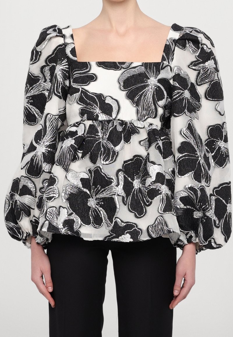 Blouse à motif floral noir et blanc avec manches longues bouffantes et encolure carrée portée avec un pantalon noir.