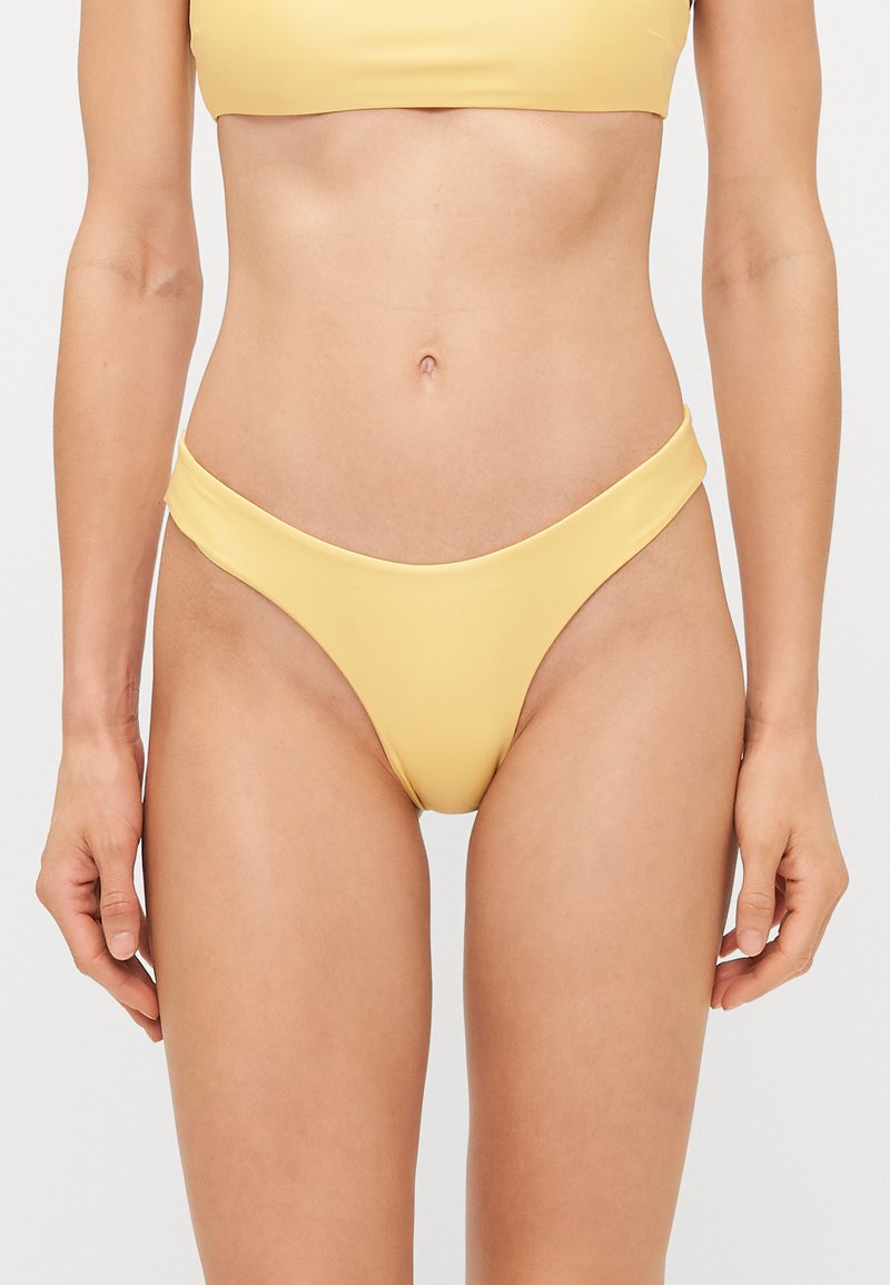 Dzelteni bikini apakšbikses, kas izgatavotas no gludas, elastīgas auduma ar zemu jostas vietu un minimālu segumu, ar elegantu izliekumu jostas vietā.