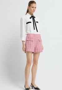Bílá blůza s černými akcenty a mašlí, doplněná o texturované růžové tweedové šortky se dvěma předními kapsami. Celkový vzhled dotvářejí černé lodičky na podpatku.