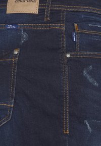 Jeans de denim oscuro con costuras amarillas, un parche de cuero en la cintura y patrones de desgaste visibles en la tela. Incluye un bolsillo para monedas.