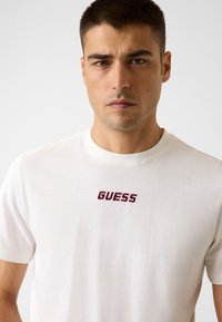 T-shirt bianca in cotone con collo rotondo e maniche corte. Logo "GUESS" ricamato in bordeaux al centro del petto. Tessuto liscio.