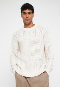 Fehér kötött pulóver, különböző fonott mintákkal és egy textúrált négyzetpanellel. Bordás, kerek nyakú és mandzsettás ujjak.