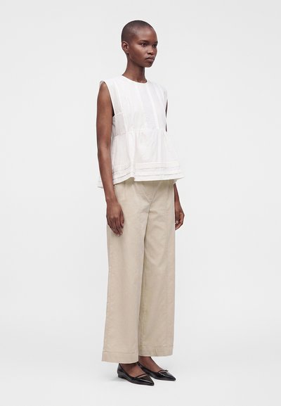 Femme aux cheveux courts portant un chemisier blanc sans manches, un pantalon beige à jambes larges et des ballerines noires pointues, debout devant un fond uni.