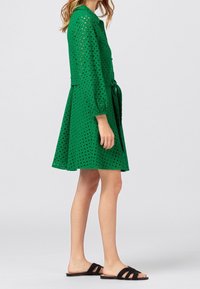 Grön klänning med ett hålmönster, långa ärmar och en utställd kjol. Kombinerad med svarta sandaler med öppen tå och ett knutet midjebälte.