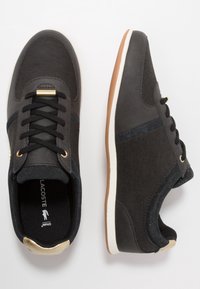 Lacoste-sneakers i mörkgrå och svarta material med en slät textur, vit mellansula och guldaccenter på snörhålen och hälen.