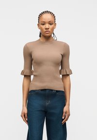 Femme aux cheveux tressés portant un haut beige ajusté en côtes avec des manches courtes à volants et un jean bleu foncé ample, debout devant un fond uni.