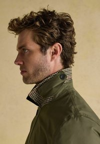 Vīrietis ar cirtainiem brūniem matiem, ģērbts olīvzaļā jakā ar paceltu pārbaudītu apkakli, skatās pa kreisi uz vienkārša bēša fona.